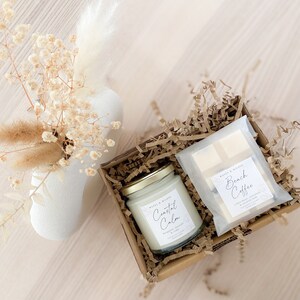 May include: A gift box filled with brown paper shreds contains a white candle with the label "Coastal Calm" and a white bar of soap with the label "Beach Coffee".