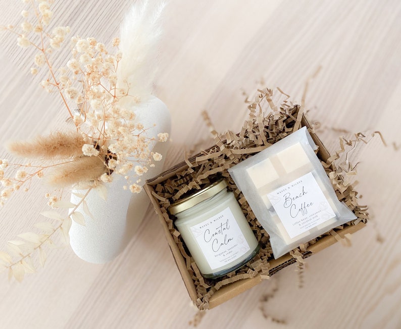 May include: A gift box filled with brown paper shreds contains a white candle with the label "Coastal Calm" and a white bar of soap with the label "Beach Coffee".
