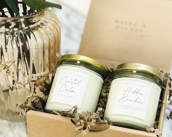 May include: Two glass candle jars with gold lids in a brown box filled with shredded paper. The candle on the left is labeled "Coastal Calm" with the scent "bergamot, lavender & cedar". The candle on the right is labeled "Hidden Beaches" with the scent "pear, midnight jasmine & whipped cream".