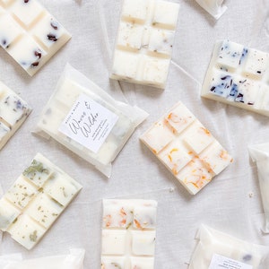 May include: Assortment of wax melts in various scents, including "Waves & Wilder" and "Sunrise Escape". Each melt is packaged in a clear plastic bag with a white label. The melts are white with various botanicals and dried flowers embedded in the wax.