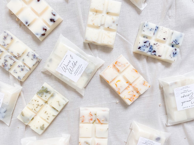 May include: Assortment of wax melts in various scents, including "Waves & Wilder" and "Sunrise Escape". Each melt is packaged in a clear plastic bag with a white label. The melts are white with various botanicals and dried flowers embedded in the wax.