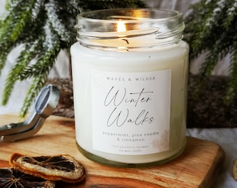 May include: A lit white soy wax candle in a glass jar with the text "Winter Walks" on a label. The candle is on a wooden board with dried orange slices and a green ribbon. The scent is peppermint, pine needle, and cinnamon.