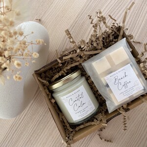 May include: A gift box filled with brown paper shreds contains a glass candle jar with a gold lid and a white label that reads "Coastal Calm" and a white bar of soap wrapped in a clear plastic bag with a label that reads "Beach Coffee".