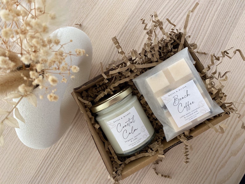 May include: A gift box filled with brown paper shreds contains a glass candle jar with a gold lid and a white label that reads "Coastal Calm" and a white bar of soap wrapped in a clear plastic bag with a label that reads "Beach Coffee".