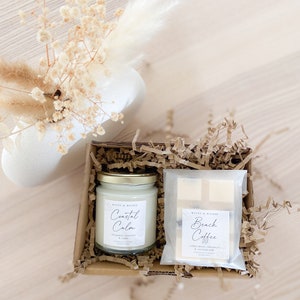 May include: A gift box filled with brown packing paper contains two candles. One candle is labeled "Coastal Calm" with the scent "Bergamot, Lavender & Cedar". The other candle is labeled "Beach Coffee" with the scent "Coffee Bean, Cinnamon & Coconut Milk".