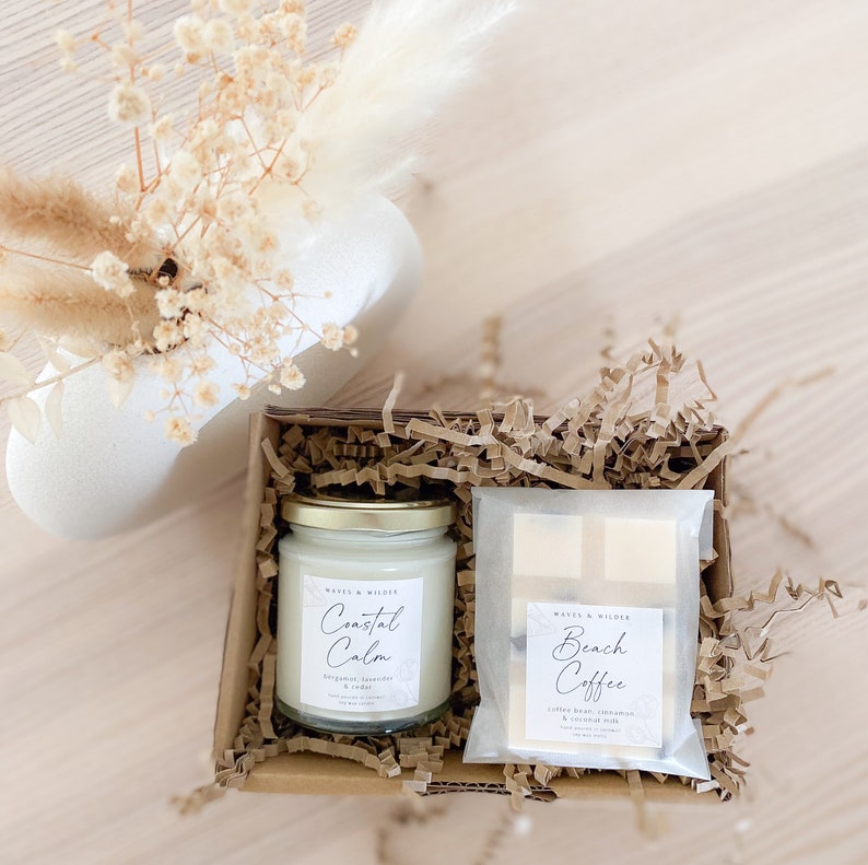 May include: A gift box filled with brown packing paper contains two candles. One candle is labeled "Coastal Calm" with the scent "Bergamot, Lavender & Cedar". The other candle is labeled "Beach Coffee" with the scent "Coffee Bean, Cinnamon & Coconut Milk".