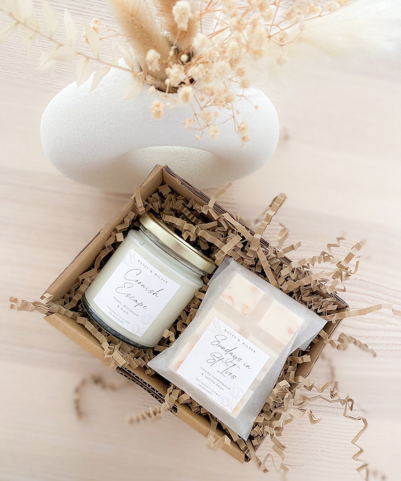 May include: A gift box filled with brown crinkle paper contains a white candle with a gold lid and a cream-colored bar of soap. The candle label reads "Cornish Escape" with the scent "Stone Driftwood & Musk". The soap label reads "Sundays in St Ives" with the scent "Creamy Sandalwood & Vanilla Bean".