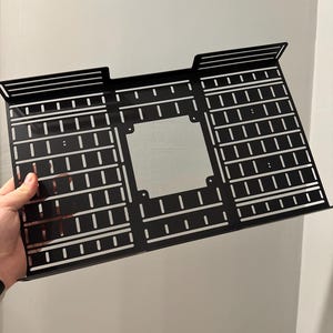 May include: A black metal computer case component with a rectangular shape and a grid-like pattern of slots and holes. The component has raised edges and a central square opening. The item is held by a hand against a white wall.