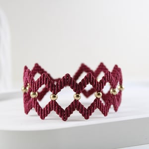 May include: A burgundy macrame bracelet with a geometric design. The bracelet features a series of diamond shapes and is accented with small, gold-colored beads. The bracelet is displayed on a white surface.