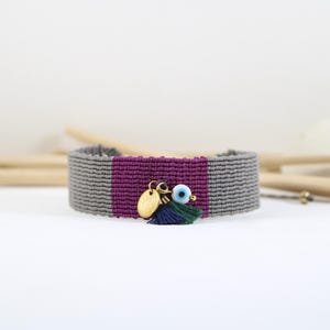 May include: A gray macrame bracelet with a central purple stripe. It features gold charms, a blue eye charm, and a small tassel. The bracelet is displayed on a white surface.