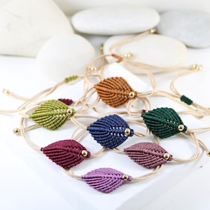 Könnte beinhalten: Handgefertigte Makramee-Armbänder mit Blättern in verschiedenen Farben, darunter Grün, Orange, Blau und Lila. Jedes Armband hat ein Blattdesign mit einer goldenen Perle und einem verstellbaren Kordelverschluss.