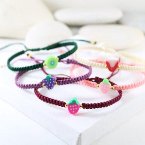 Peut inclure: Plusieurs bracelets d'amitié colorés avec des breloques en forme de fruits. Les bracelets présentent des couleurs variées, notamment vert, violet, rose et bordeaux, avec des détails dorés. Les breloques représentent un kiwi, une fraise et un raisin.