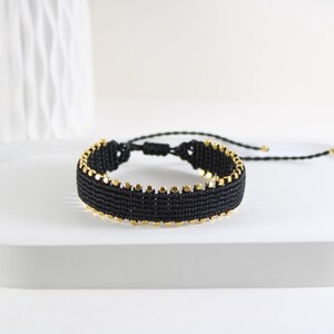 Peut inclure: Bracelet en macramé noir avec des accents dorés. Le bracelet est fait de fils noirs tissés et comporte de petites perles dorées espacées régulièrement sur les bords. Le bracelet est présenté sur un plateau blanc.