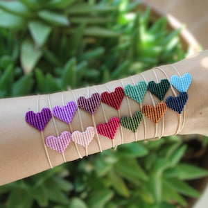 Könnte beinhalten: Eine Sammlung von Makramee-Armbändern mit kleinen herzförmigen Anhängern in verschiedenen Farben wie Lila, Pink, Rot, Grün, Orange und Blau, die jeweils an einer dünnen, neutralfarbenen Kordel um das Handgelenk getragen werden.