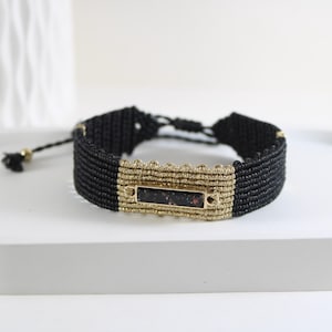 Peut inclure: Bracelet en macramé noir avec une pièce maîtresse rectangulaire dorée. La pièce maîtresse a une pierre sombre mouchetée. Le bracelet est réglable avec un cordon noir et une perle dorée.