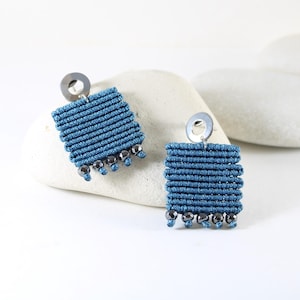 May include: A pair of square, blue macramé earrings with silver-tone metal accents. Each earring features a square design with a series of horizontal lines and small, dangling beads. The earrings are displayed on a white stone against a white background.