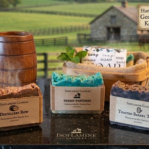 Puede incluir: Una disposición de jabones hechos a mano y un barril de madera. Los jabones están etiquetados como "Distillery Row", "Green Pastures" y "Toasted Barrel Toffee". Una cesta contiene jabones adicionales y ramitas de menta. El fondo muestra un paisaje rural.