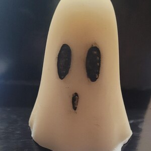 May include: A cream-coloured ghost-shaped candle with black oval eyes and a small open mouth. The candle has a smooth texture and a slightly rounded top, ideal for Halloween or spooky decor.