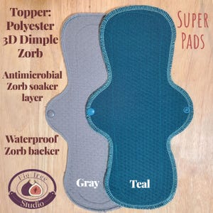 May include: Two reusable cloth menstrual pads in gray and teal. The gray pad has a snap closure and is labeled "Gray." The teal pad, also with a snap, is labeled "Teal." The pads are made with a polyester topper, Zorb layers, and a waterproof backer. The words "SUPER PADS" are in the upper right corner.