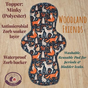 May include: A reusable menstrual pad with a woodland animal print featuring foxes, deer, and rabbits. The pad has a dark blue background and is labeled "Woodland Friends." It is washable and designed for periods and bladder leaks.