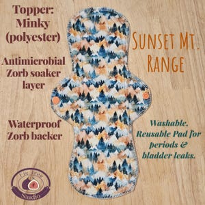 May include: A reusable menstrual pad with a mountain range pattern in blue, orange, and white. The pad has a minky polyester topper, an antimicrobial Zorb soaker layer, and a waterproof Zorb backer. The text "Sunset Mt. Range" is visible.