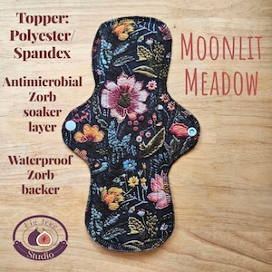May include: A reusable menstrual pad with a floral design. The pad features a black background with colorful flowers and foliage. The text "MOONLIT MEADOW" is displayed in red. The pad has snap closures and is made with polyester, spandex, and Zorb.