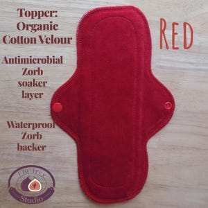 May include: A red, reusable cloth menstrual pad with a rounded shape and snap closures. The pad is made with organic cotton velour, an antimicrobial Zorb soaker layer, and a waterproof Zorb backer. Text on the pad reads "RED".
