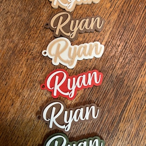 May include: Six personalized name tags with the name "Ryan" written in a cursive font. The tags are made of different materials and colors, including brown, beige, white, red, and green.