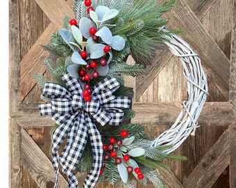 May include: A white wicker wreath decorated with a black and white checkered ribbon bow, red berries, and green pine sprigs.