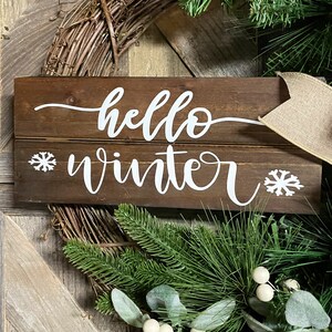 May include: A wooden sign with the text "hello winter" in white cursive lettering. The sign is decorated with two white snowflake designs. The sign is surrounded by a green wreath with white berries.