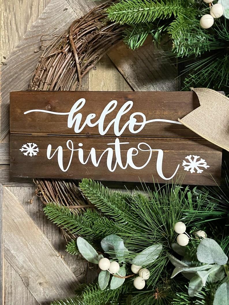 May include: A wooden sign with the text "hello winter" in white cursive lettering. The sign is decorated with two white snowflake designs. The sign is surrounded by a green wreath with white berries.