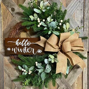 May include: A rustic wreath with a burlap bow and a wooden sign that says "hello winter". The wreath is decorated with greenery, white berries, and eucalyptus.