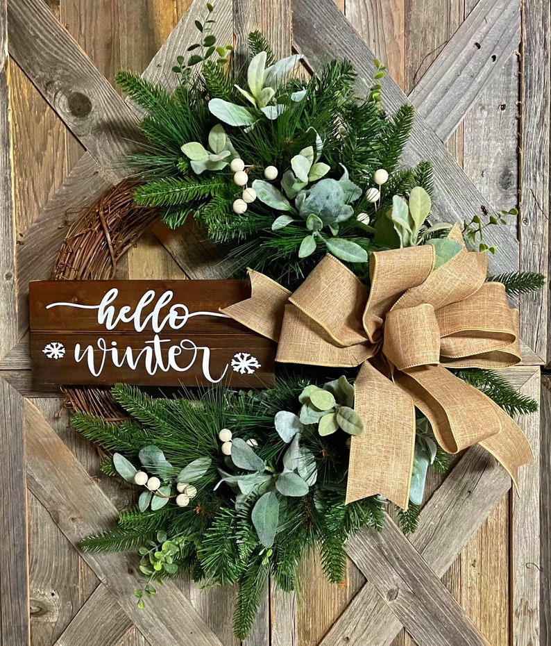 May include: A rustic wreath with a burlap bow and a wooden sign that says "hello winter". The wreath is decorated with greenery, white berries, and eucalyptus.