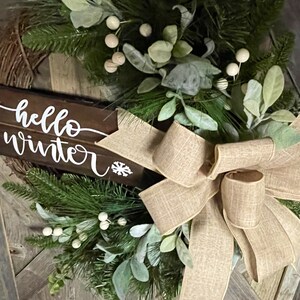 May include: A rustic wooden sign with the text "hello winter" in white lettering, decorated with a burlap bow and greenery with white berries.
