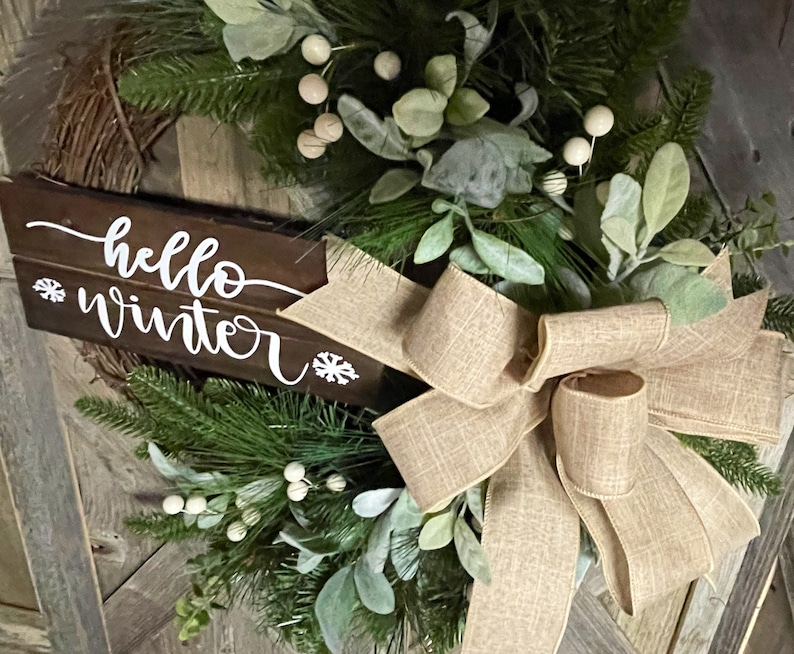 May include: A rustic wooden sign with the text "hello winter" in white lettering, decorated with a burlap bow and greenery with white berries.