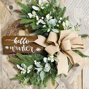 May include: A rustic winter wreath with a burlap bow, green pine branches, white berries, and a wooden sign that says "hello winter".