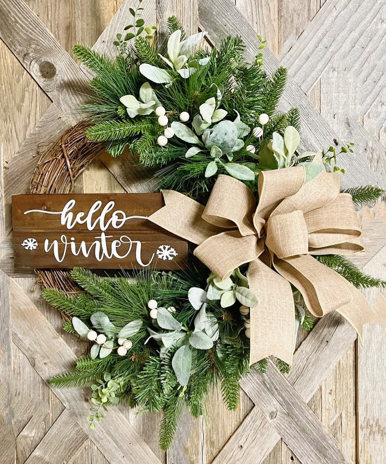 May include: A rustic winter wreath with a burlap bow, green pine branches, white berries, and a wooden sign that says "hello winter".