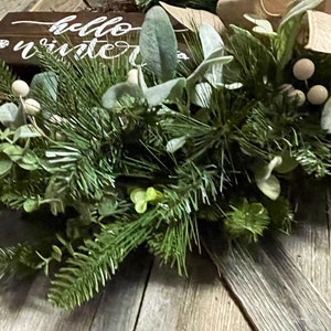 May include: A green and white winter wreath with a wooden sign that says "hello winter".