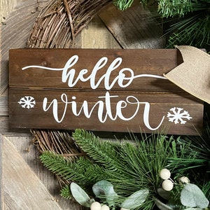 May include: A wooden sign with the text "hello winter" in white cursive lettering. The sign is decorated with two white snowflake designs. The sign is surrounded by a green wreath with white berries.