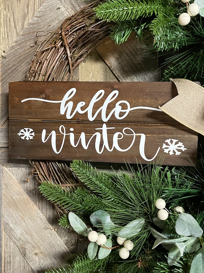 May include: A wooden sign with the text "hello winter" in white cursive lettering. The sign is decorated with two white snowflake designs. The sign is surrounded by a green wreath with white berries.