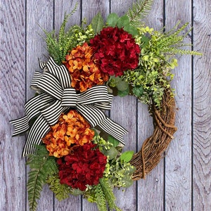 May include: A wreath made of grapevine with a black and white striped bow. The wreath is decorated with red, orange, and green hydrangeas and greenery.