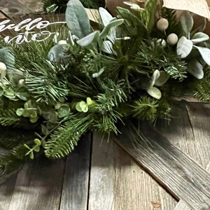 May include: A wooden sign with the text "Hello Winter" on it, decorated with a green garland of pine branches, eucalyptus leaves, and white berries. The garland is placed on a rustic wooden background.
