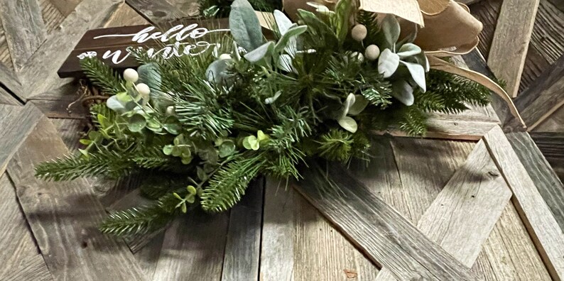 May include: A wooden sign with the text "Hello Winter" on it, decorated with a green garland of pine branches, eucalyptus leaves, and white berries. The garland is placed on a rustic wooden background.