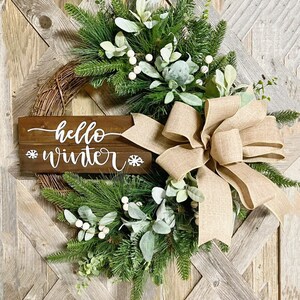 May include: A rustic wooden sign with the text "hello winter" and snowflakes on either side, decorated with a burlap bow and a wreath of pine branches, eucalyptus, and white berries.