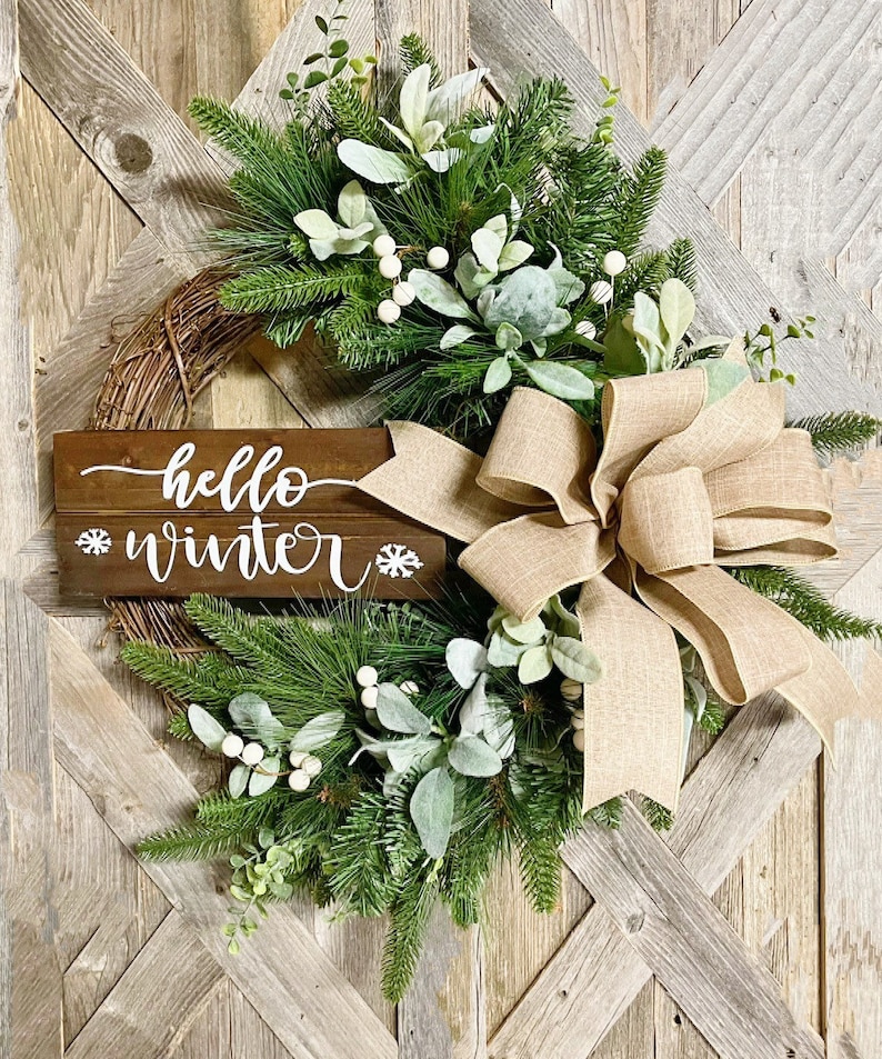 May include: A rustic wooden sign with the text "hello winter" and snowflakes on either side, decorated with a burlap bow and a wreath of pine branches, eucalyptus, and white berries.