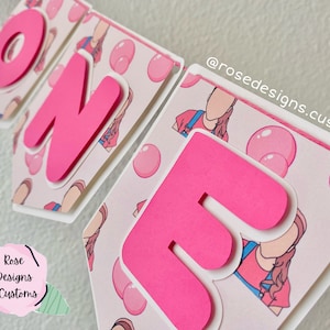 May include: Pink and white banner with the word "ONE" in large pink letters. The banner has a repeating pattern of pink bubbles and a cartoon girl wearing a pink shirt and blue overalls. The banner also includes the text "@rosedesigns.customs".