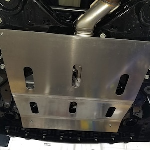 Puede incluir: Un escudo de protección de bajos de carrocería de metal plateado con múltiples recortes para acceder a los componentes del vehículo. El escudo está fijado a la parte inferior de un coche con tornillos.