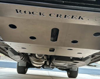 May include: A silver-colored metal skid plate installed under a vehicle, featuring the laser-cut text ROCK CREEK and two mountain icons. The plate has a smooth, brushed finish with several circular and elongated ventilation cutouts throughout.