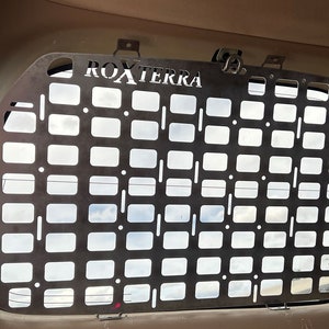 May include: A metal panel with a grid pattern and the text "ROXTERRA" cut out at the top. The panel is installed in a vehicle and has several mounting points. The grid design allows for visibility and potential storage options.