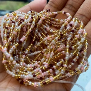 May include: A collection of beaded waist beads in a hand. The beads are a mix of gold, pink, white, and dark red. The beads are strung together on a thin, white string. The beads are small and round.